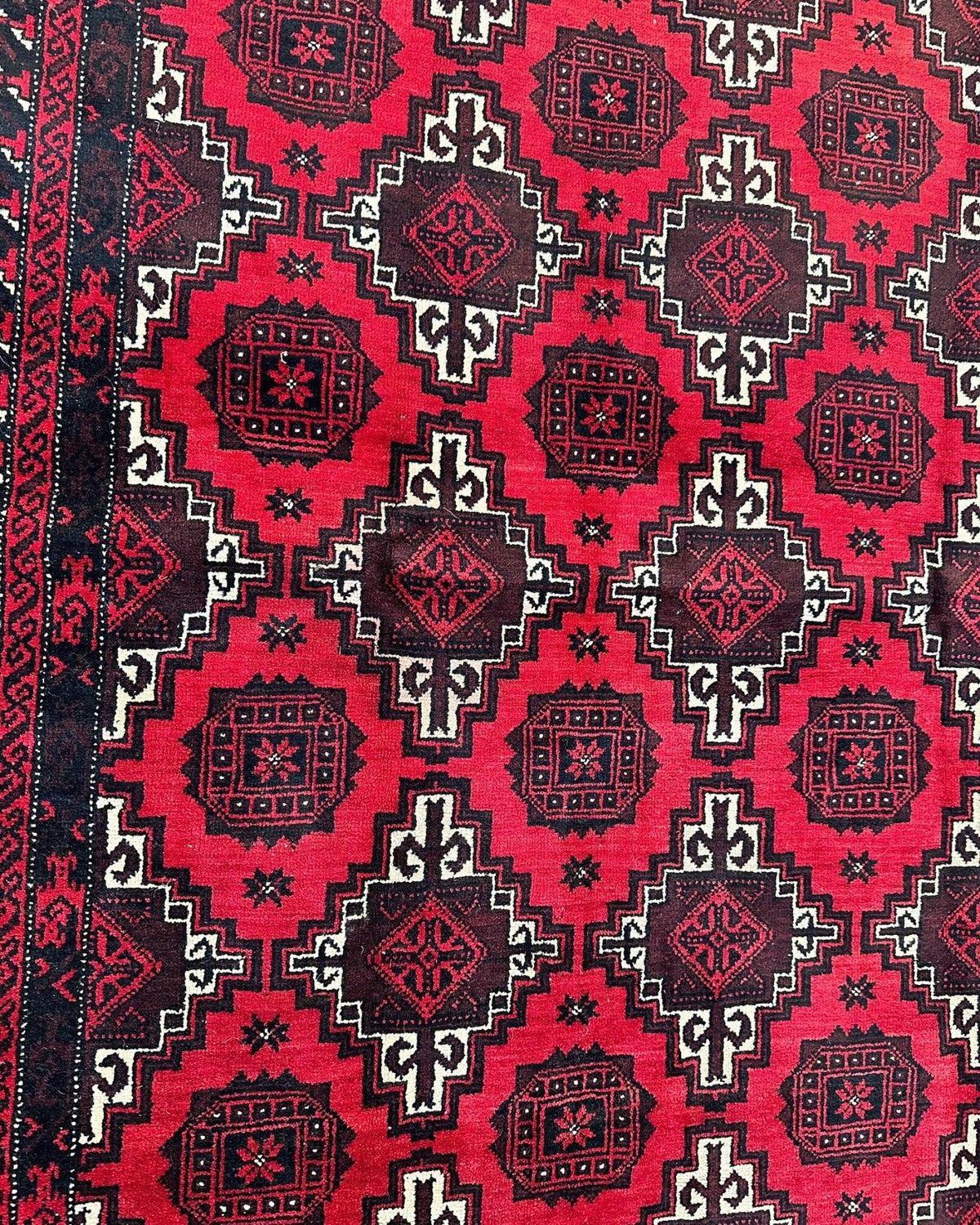 Close-up of a hand-knotted Afghani Baluch wool rug with repeating red and black geometric medallions framed by a patterned border.