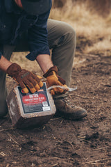 Person wearing brown gloves and khaki pants opening a plastic-wrapped block labeled 'BIG TINE FORTIFIED DEER BLOCK' while kneeling on dirt ground outdoors.