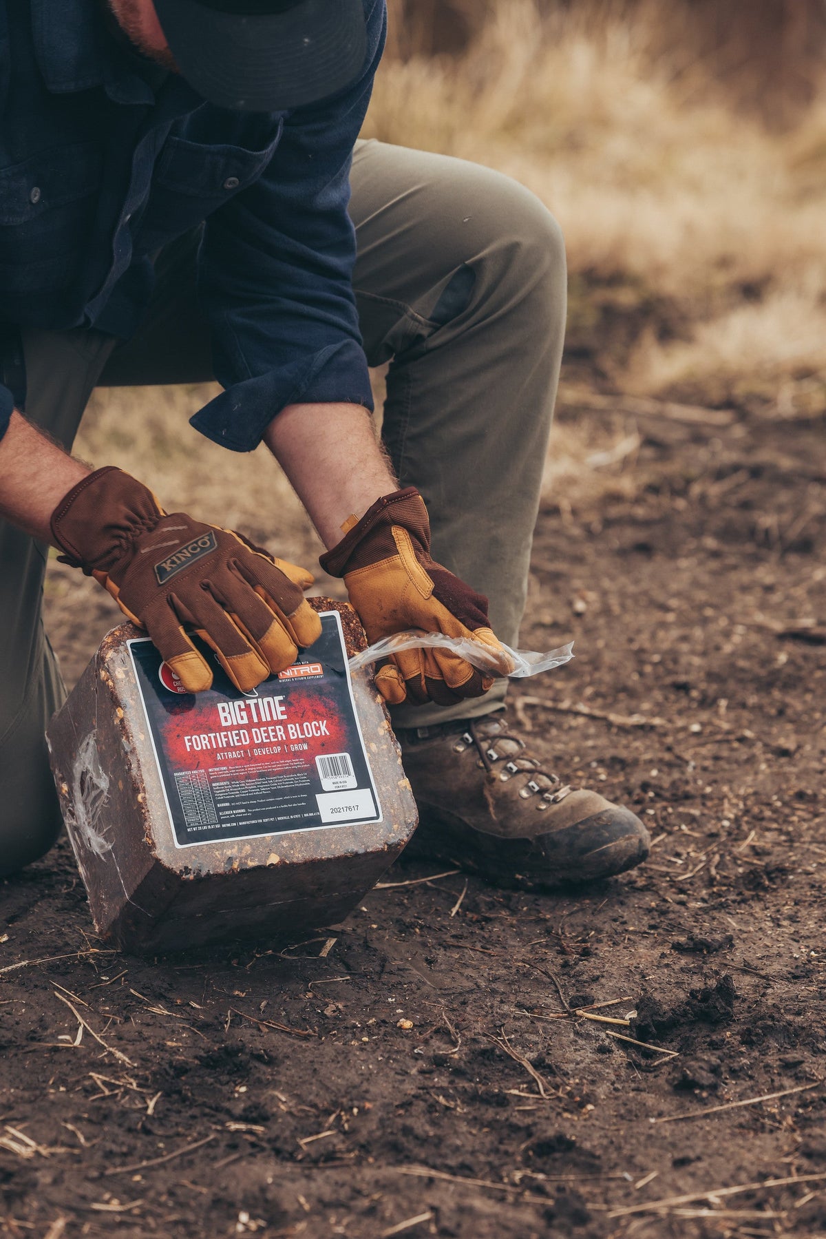 Person wearing brown gloves and khaki pants opening a plastic-wrapped block labeled 'BIG TINE FORTIFIED DEER BLOCK' while kneeling on dirt ground outdoors.
