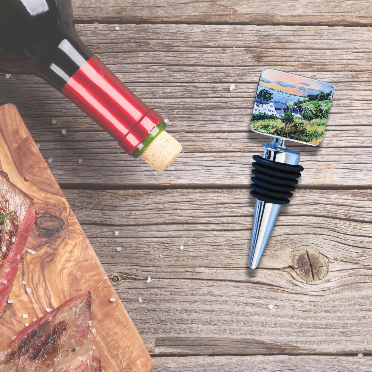 Wine bottle stopper with a painted square top depicting a sunset lake view and houses, lying on a weathered wooden table next to a wine bottle and a cutting board.