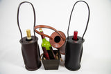 Two cylindrical leather wine totes in dark brown and black with stitched edges hold a wine bottle each; a brown leather strap is draped over a small basket with a green bottle inside.