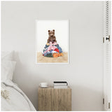 Framed canvas art on a white wall showing a brown bear cub sitting atop a colorful pile of laundry, above a wooden bedside table with books and a cup beside a white bed.