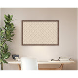 Wall-mounted beige pinboard with a subtle green lattice pattern framed in dark wood above a light wooden desk with books and a green leafy plant to the left.