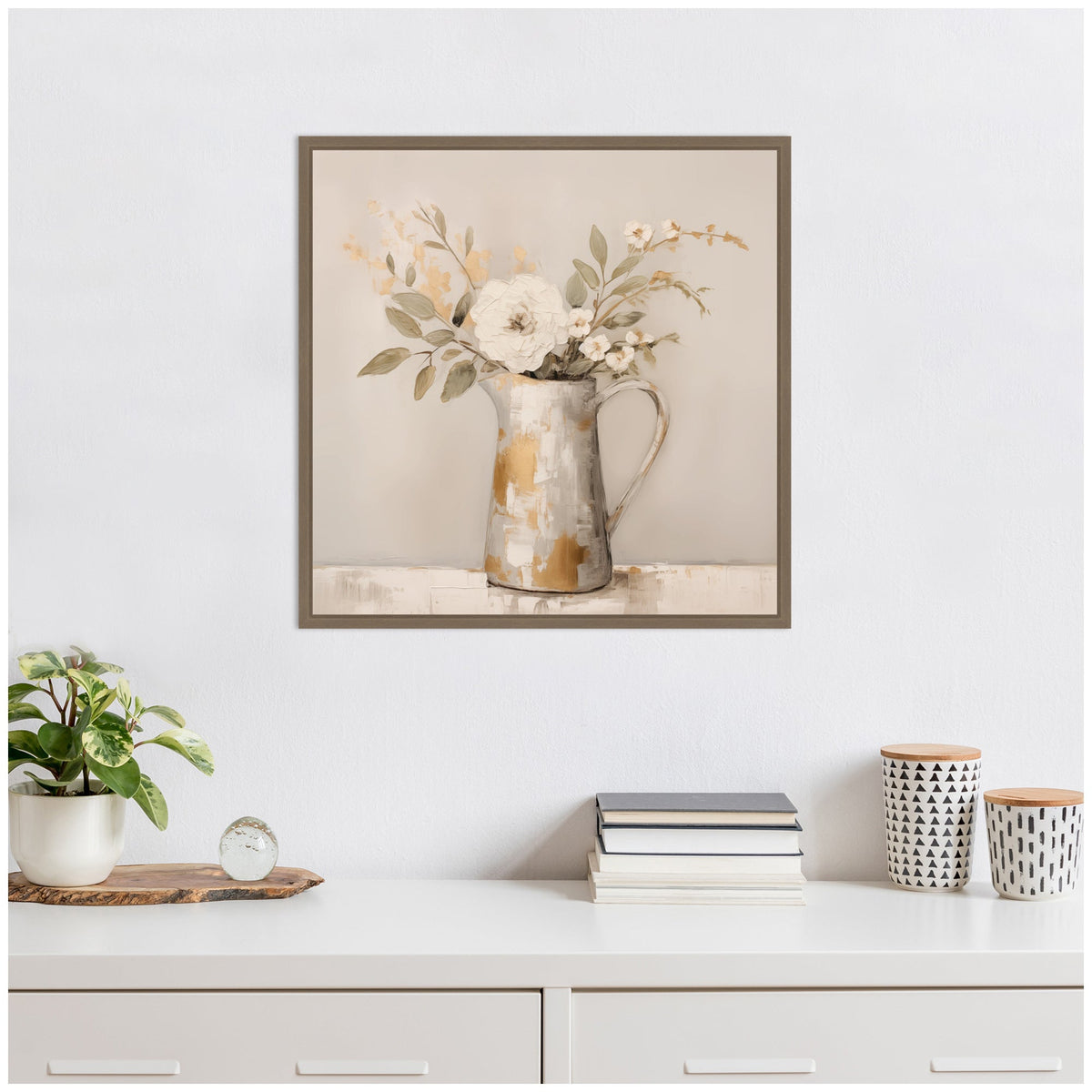 Square framed artwork of a rustic pitcher holding cream and beige flowers with green leaves, hanging above a white cabinet with books, plant, and patterned containers.