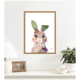 Framed watercolor and mixed media portrait of a bunny with large black eyes, green leafy ears, and colorful flowers on its head and chest, hanging on a white wall above a shelf with decor.