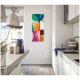 Narrow vertical framed canvas with colorful abstract geometric shapes including green, blue, yellow, orange, pink, and red, hanging on a white kitchen wall between two countertops.
