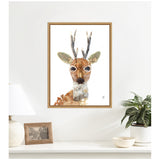 Framed canvas print of a stylized buck with large eyes, textured antlers, and earthy tones, hanging centered on a white wall above a shelf with decor.