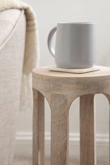 Light wood round drink table with four legs featuring subtle curved cutouts, holding a gray ceramic mug on a square coaster beside a beige upholstered chair.