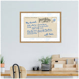 Framed canvas showing a vintage-style postcard with blue handwritten text and seashell stamp, hanging above a wooden desk with plants and office supplies.