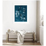 Framed aerial photo of a school of humpback whales swimming in deep blue ocean water, displayed above a simple wooden bench with a draped cloth and open book.