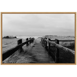Black and white photograph of a wooden boardwalk flanked by grassy dunes leading to a cloudy beach horizon, framed in light wood.