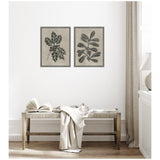 Two framed black and beige botanical prints featuring holly leaves with berries and broad leaves, hung side by side above a wooden bench with a white blanket and book.