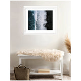 Framed aerial photograph of dark black sand beach with white ocean waves crashing along the shoreline, hanging above a white bench with a beige fur throw and a brown bag nearby.