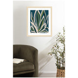 Abstract botanical print with layered, elongated leaves in green, beige, and blue tones on a deep teal background, framed in light wood and hung above a dark olive chair and potted plant.