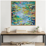 Abstract painting of a water lily pond with green, blue, yellow, and pink brushstrokes in a wooden frame hanging above a wooden console table with books, a cup, and a white vase.