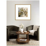 Framed wall art print of an olive branch with green olives and leaves, set above a round wooden table with flowers, between two dark gray armchairs in a neutral room.