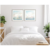 Two framed abstract landscape prints above a white bed, featuring soft pastel blues, pinks, and yellows suggesting sky and water scenes.