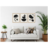 Three framed black geometric abstract prints with simple shapes on white walls above a white cushioned sofa with patterned and neutral pillows in a modern living room with green plants.