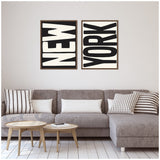 Two black and white framed canvases with bold, large text reading 'NEW' and 'YORK' displayed side-by-side above a modern gray sectional sofa with striped and solid cushions.