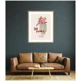 Framed art print titled 'Les Fleurs' featuring a minimalist line drawing of flowers in a striped vase with red handwritten text, hung above a brown leather sofa.