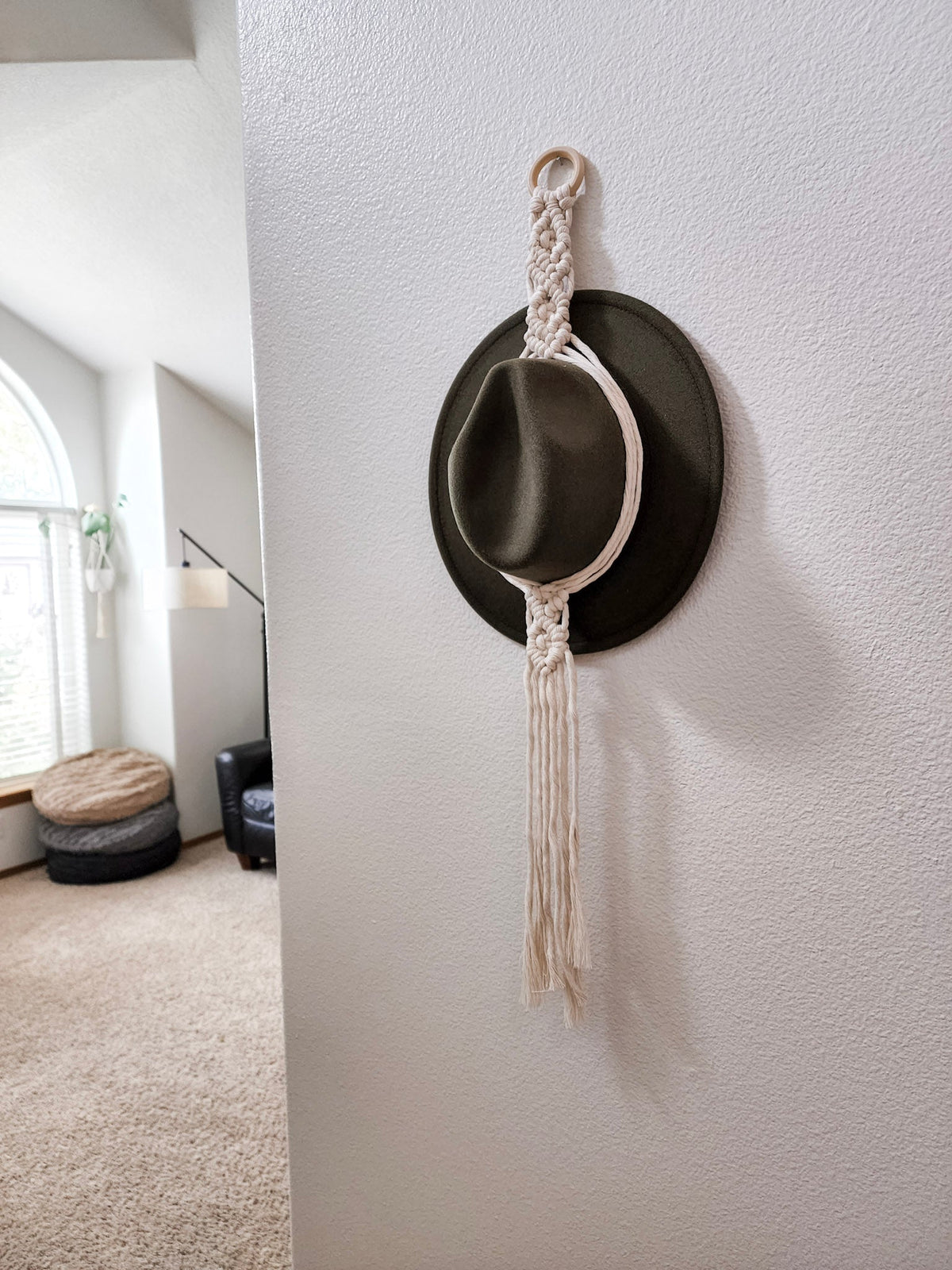 White macrame rope hanger with fringe suspending a dark olive-green felt hat, mounted on a white wall near a living space with arched window and neutral decor.