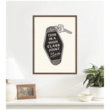 Minimalist black and white framed artwork of a vintage-style keychain with text that reads 'This is a high class joint, act accordingly' hanging on a white wall above a clean white surface with a plant, lamp, and photo frame.