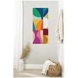 Tall vertical abstract wall art with colorful geometric shapes in green, blue, yellow, pink, red, and white framed on light gray wall near woven basket and white vase with pampas grass.