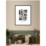 Abstract black leaf and petal shapes on beige background in a black frame above green wainscoting and a wooden bench with pillows and a draped blanket.