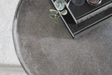 Close-up of round coffee table with textured dark gray surface, displaying the edge and top with stacked books and a small green plant on top.