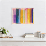 Abstract framed canvas featuring vertical vibrant lines in purple, blue, yellow, orange, and pink shades hung on a white wall above a white cabinet with books, plants, and patterned containers.