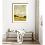 Abstract landscape painting with soft green and orange hues framed in light wood hanging above a wooden bench with a folded blanket and open book in a neutral-toned room.