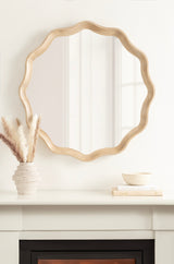 Round wall mirror with a scalloped, light wood frame hanging above a white mantel displaying a textured beige vase with pampas grass and stacked books with a small bowl.