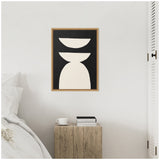 Geometric abstract framed canvas with three white shapes on black background, hung above a rustic wood bedside table holding books and a white cup.