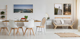 Bright modern dining and living area with white walls, wooden table and chairs, beige sofa, and framed nature photographs above the sofa and on the far wall.