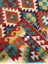 Close-up of Afghani Maimana Kilim wool rug with geometric patterns in red, orange, yellow, teal, and white, showing fringed edge and intricate diamond shapes.