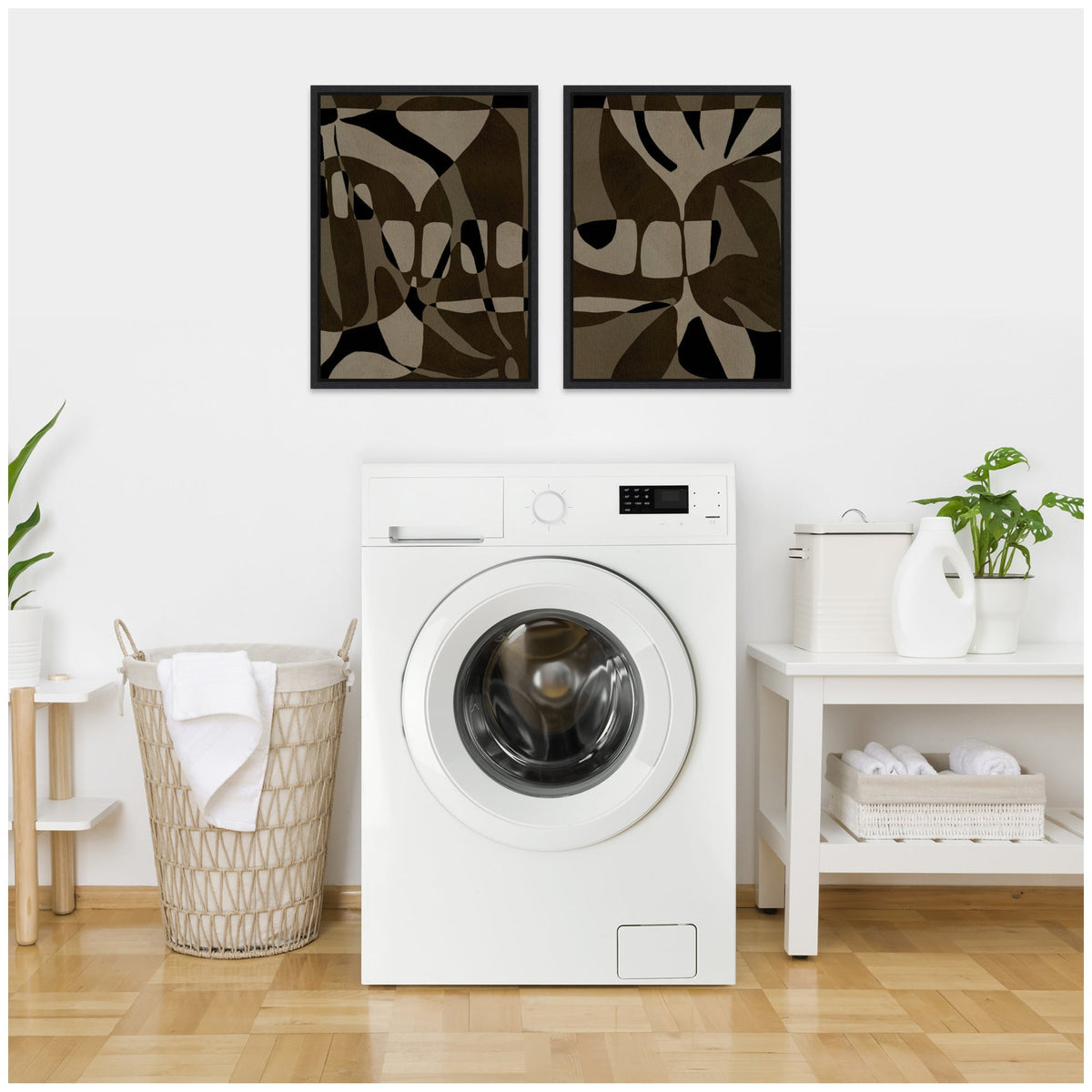Two framed abstract canvas art pieces with black, brown, and beige organic shapes paired above a white front-loading washing machine in a minimalist laundry room.