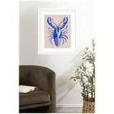 Framed blue lobster illustration on beige background hanging above dark green upholstered chair and potted olive tree in woven basket.