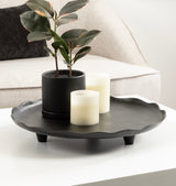 Black round decorative tray with scalloped edges on three short legs holding a small potted plant with variegated leaves and two white pillar candles, on a white table.