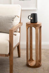 Tall cylindrical wooden stool with vertical cutouts and round top holding a black mug, positioned next to a beige upholstered chair with wooden frame and gray pillow.