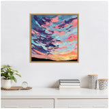 Framed canvas artwork depicting a vivid sunset with swirling clouds in shades of blue, pink, and orange above a dark horizon, hanging on a white wall above a minimalist white desk with books, patterned containers, and a plant.