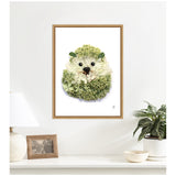 Framed print of a hedgehog composed of green and white flower petals and leaves on a white wall above a white surface with a lamp, photo frame, and potted plant.