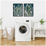 Two framed botanical prints with green and white leaves on dark blue backgrounds hang above a white front-loading washing machine in a bright laundry room with a wicker basket and white shelving.