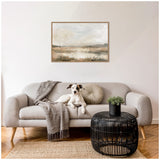 Neutral-toned abstract landscape painting with soft beige, gray, and white hues, framed and hanging above a light gray sofa with a dog, throw, round cushion, and black wicker coffee table.