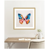 Square framed artwork of a colorful butterfly with bold brushstroke wings in blue, orange, and pink hues on a light background, hanging above a rustic wooden desk.