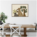 Framed canvas wall art depicting three rustic vases with green plants on a beige background, hanging above a wooden dining table with white chairs and cushions.