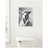 Black and white framed portrait of a llama wearing a bathrobe, hanging above a white toilet tank against a plain wall near a window with blinds.