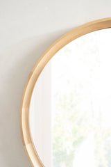 Close-up of a round wall mirror with a smooth, segmented light wood frame against a pale wall, softly reflecting a blurred window and greenery.