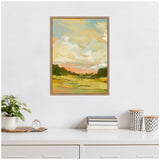 Framed canvas painting of a landscape with green fields, dark tree line, and a cloudy sky in soft blues, yellows, and pinks hanging above a white desk with books and patterned containers.