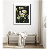 Framed artwork featuring white flowers with green leaves on a dark background hanging above a wooden bench with a light-colored throw and an open book in a softly lit room.