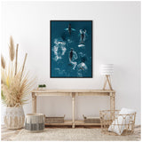 Framed aerial photo of five humpback whales swimming in blue ocean water with white splashes, hanging above a rustic wooden console table in a bright room.